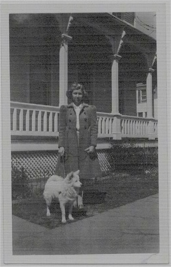 The front right side of a white American Eskimo dog that is standing out in front of a house with a lady in 1941