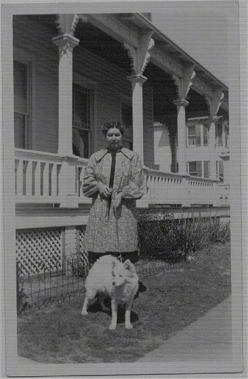 A 1941 photograph of a white American Eskimo dog that is standing out in front of a house looking to the left. There is a lady standing behind it.
