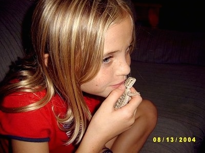 A blonde haired girl has a baby Bearded Dragon in her hand and she is holding it close to her mouth.