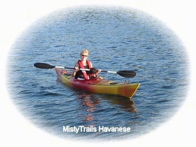 A lady is sitting behind a black with white Havanese in a Kayak out in the middle of open water.