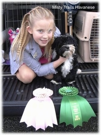 A blonde haired girl is sitting on a truck bed holding onto a black and white puppies neck to pose it. They have a pink ribbon and a green ribbon in front of them