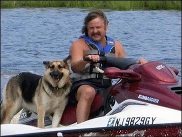 A German Shepherd is riding on a Jet Ski with a Man in a blue life jacket