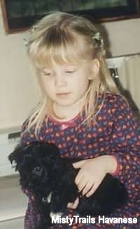 A little blonde haired girl is holding a black with white Havanese puppy in her hands