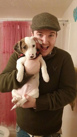 Bernard the white with tan and black Doxle Puppy is wearing a red and black plaid shirt. He is being held up in the air by a man in a green hat.
