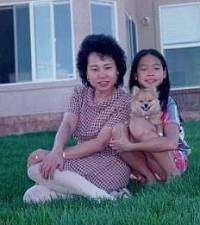 A mother and a daughter are sitting in front of a house. A tan with white Pomeranian puppy is sitting in the daughters lap.