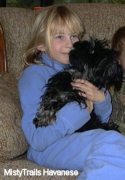A smiling blonde haired girl in blue has a black Havanese puppy in her arms sitting on a tan couch.