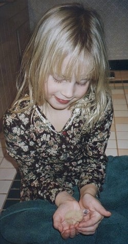 Close up - A blonde haired girl is sitting on a tiled floor and she is cupping her hands to to hold a newborn Cockatiel bird.
