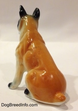The back left side of a red and white with black bone china Boxer figurine that is in a sitting pose. The figurine has a short tail.