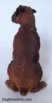 The back of a brown with black and white Boxer figurine that is in a sitting pose and it is made out of resin. The tail of the figurine is short.