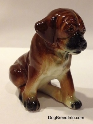 The front right side of a brown with black and white ceramic Boxer puppy figurine with uncropped ears in a sitting pose. The figurine has a black muzzle and ears that hang down to the sides.