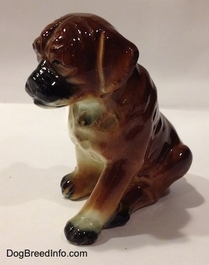The front left side of a brown with black and white ceramic Boxer puppy figurine with uncropped ears in a sitting pose. The figurine has tiny black eyes.