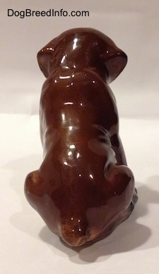 The back of a brown with black and white ceramic Boxer puppy figurine with uncropped ears in a sitting pose. The figurine is glossy.