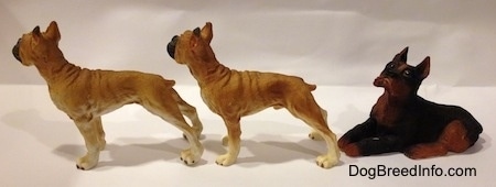The left side of two tan with white and black Boxer standing figurines are in front of a black with brown laying Boxer figurine.