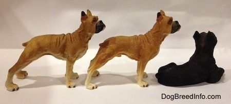 The right side of two tan with white and black Boxer standing figurines are behind a black with brown laying Boxer figurine.