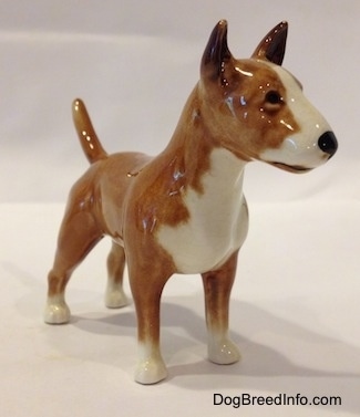 The front right side of a brown with white Bull Terrier figurine. The figurine has black circles for eyes and a nose. The dog has a wide muzzle with a shallow stop and a black nose. It has white down the center of its snout.