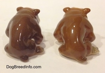 The back side of two brown with white miniature Bulldogs that are in a sitting pose. The figurines have short tails.