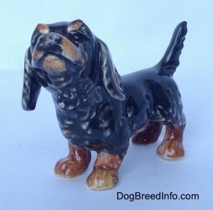 The front left side of a figurine of a black and brown Dachshund in a standing pose with its tail up. The figurine has a lot of detail in its face.