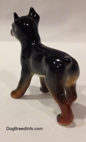 The back left side of a figurine that is a black and brown Doberman Pinscher puppy figurine. The figurine has a glossy body.