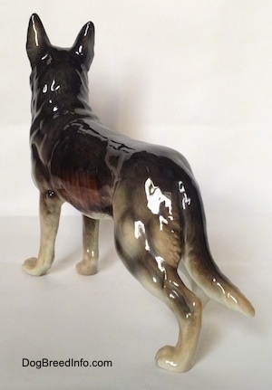 The back left side of a figurine of a standing German Shepherd. The figurine has a long tail that is the length of its leg.
