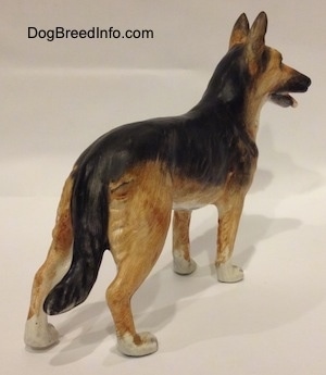 The back right side of a black and tan with white porcelain German Shepherd standing figurine. The figurines tail is long.