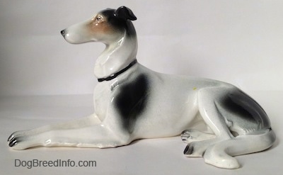 The left side of a white with black and tan Greyhound figurine in a lying down pose. The figurine has a couple of large black spots on its body.