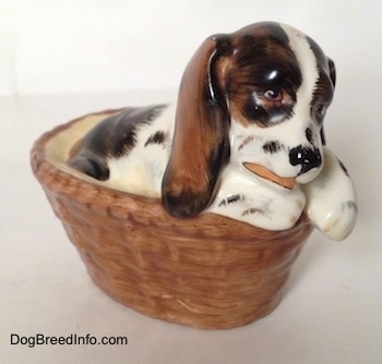 The front right side of a white with brown and black Russian Spaniel puppy in a basket figurine. The figurine is painted to look like it is licking its paw.