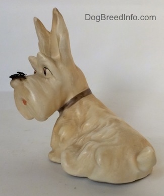 The back left side of a white and cream figurine of a Scottish Terrier with a fly on its nose. The figurine has fine hair details along its back.