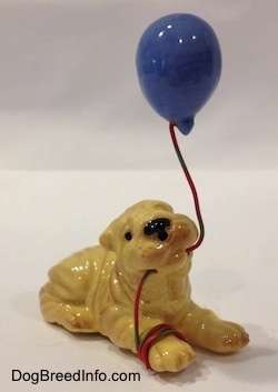 The front right side of a figurine of a Shar-Pei lying with a blue balloon in its mouth. The figurine has black circles for eyes.