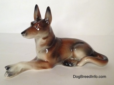 The left side of a brown and white with black German Shepherd figurine lying. The figurine is glossy.