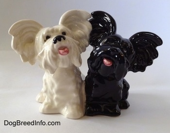A figurine of a black Skye Terrier and a white Skye Terrier. The figurines mouths are painted open and they are looking up.