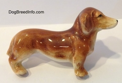 The right side of a tan ceramic Dachshund dog figurine. The figurine has a broken leg. The ear of the figurine is attached to its body and it is hard to differentiate.