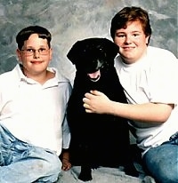 Two boys are sitting around a black Labrador Retriever that is sitting on a backdrop. The dogs mouth is open and it looks like it is smiling
