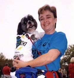 A lady in a blue shirt is holding a black with white Shih Tzu dog against her shoulder. The Shih Tzu and the lady are both looking forward.