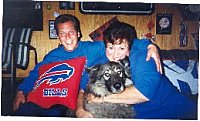 A man has his arm around the shoulder of a lady, who has her arms around a wolf-looking, black, grey with white mixed breed dog. They are both wearing blue sitting on a couch in front of a wood wall in a home. The man has a red and blue sports team pillow on his lap.