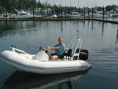 A black Schipperke dog is standing on the back of a boat that is on the water. There is a man sitting in the drivers seat of the boat.