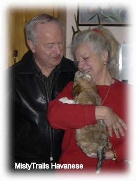 A lady in a red long sleeve shirt is holding a tan with white Havanese puppy against her body and next to her is a guy wearing a black leather jacket.