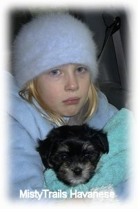 Close up - A sad looking girl in a fuzzy blue hat and a big blue jacket is holding a black with gray Havanese puppy that is wrapped in a towel.