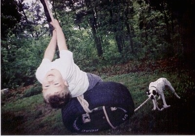 The backside of a boy that is swinging on a tire swing and looking back. There is a white with black dog pulling a rope attached to the tire. 