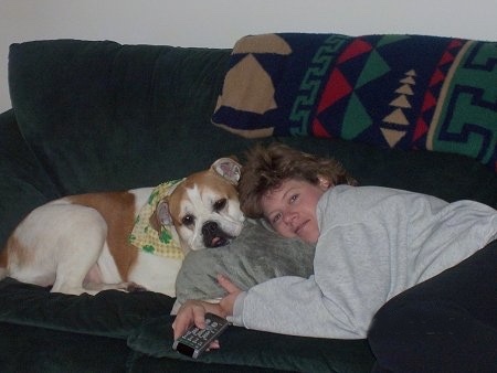 A tan with white Olde English Bulldogge wearing a yellow and green bandana laying on a green couch with its head on a gray pillow. The dog is head to head with a lady in a gray sweatshirt who is also laying on the pillow and holding a TV remote control.