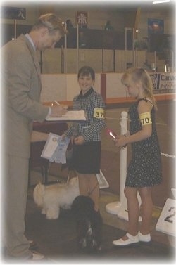 A blonde-haired girl is standing behind a black with white dog and to she is being judged by a blonde-haired man in a tan suit. Next to the girl is a girl in a black and white jacket holding the leash of a white dog and smiling.
