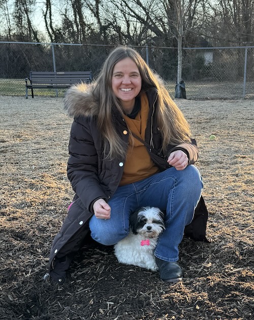 A long haired girl kneeling down with her little doodle dog at a dog park