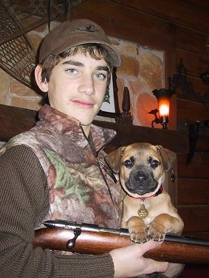 Ace the Black Mouth Cur puppy being held by a male person who is posing with a gun