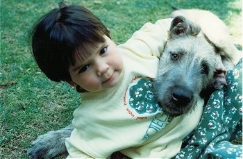 A child has its arm around the head of a white with tan Irish Wolfhound that is laying in grass