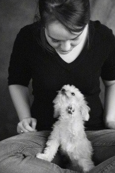A black and white photo of Ellie the Chorkie sitting in her owners lap who is sitting in indian style. Ellie's owner is looking down at her and Ellie is looking up at the woman. One of Ellie's paws is in the air
