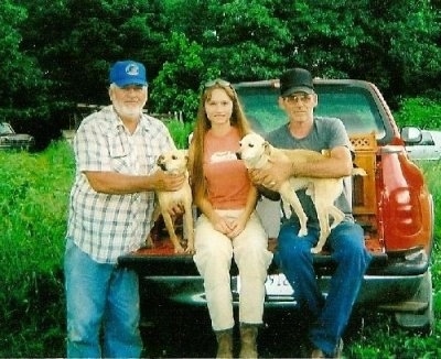 Three people at the back of a red pick up truck with an open tail-gate. Two men with a lady in the middle. The men are wearing baseball type caps and blue jeans and are holding tan and white medium-sized hunting dogs.