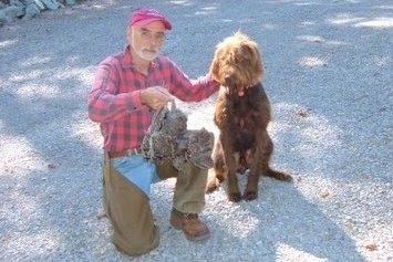 A brown wavy-coated, Pudelpointer dog is sitting on a rocky surface next to a kneeling man in a red cap, red and black flannel shirt, tan covered blue jeans and brown boots. The man is holding a dead bird in its left hand and he is touching the dog with his right hand