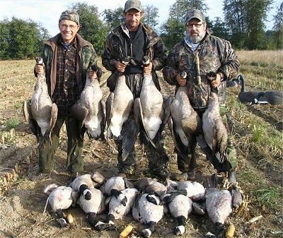 Three men are holding 6 dead geese. A goose in each hand. In front of them is 10 more dead Geese laying on the ground.