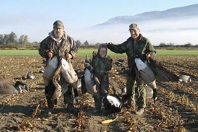 Two men holding dead Geese are standing around a boy holding a dead goose. One man has his hand on the head of the boy