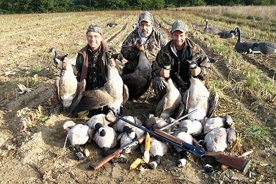 Three men are holding a dead goose in each hand kneeling behind a line of dead geese with two guns over top of each other making an X on top of the pile of dead geese on the ground.