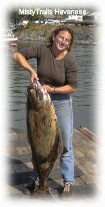A lady is standing on a dock and she is lifting up a Halibut fish. She is looking forward.